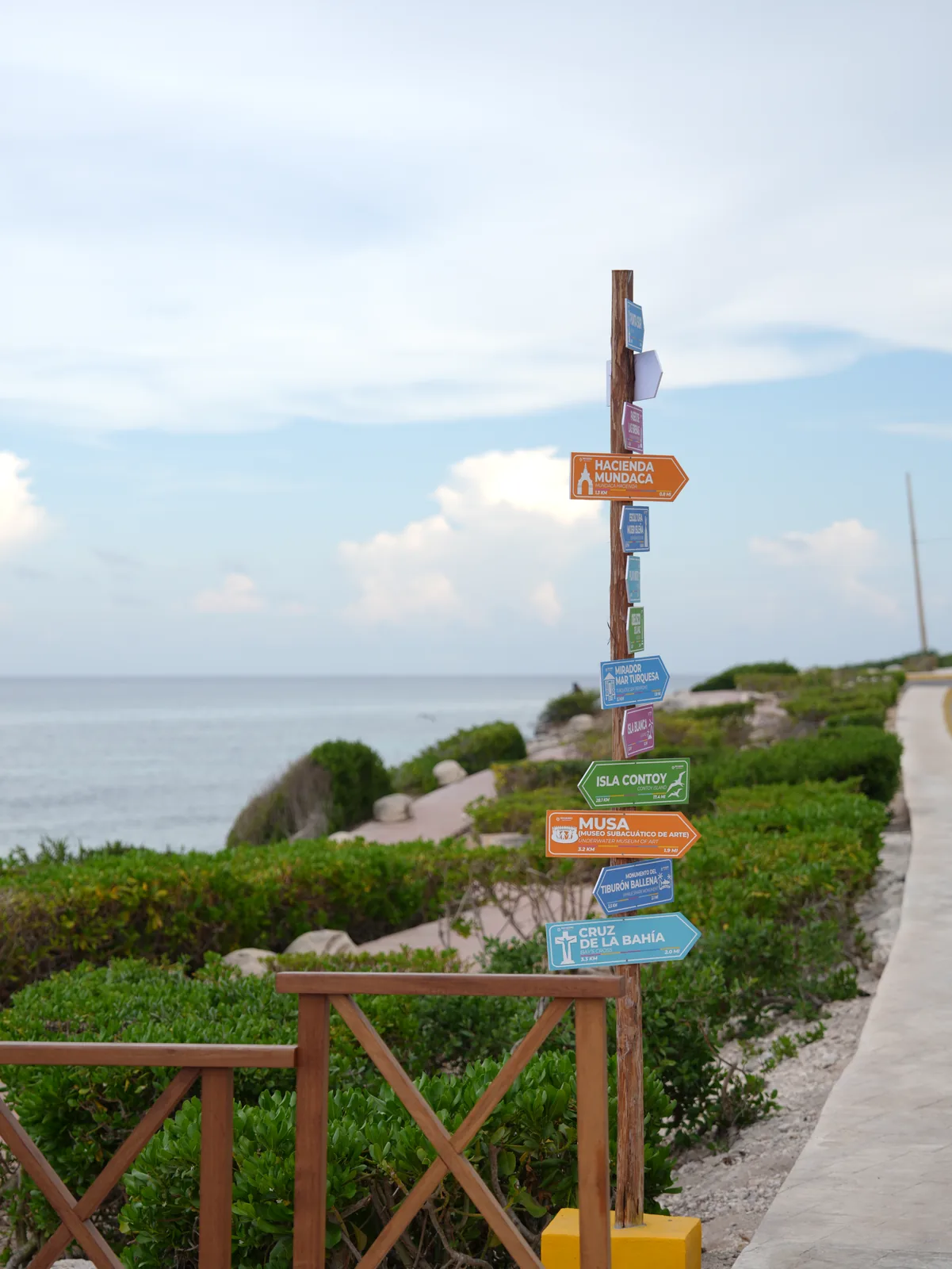 A wooden signpost with colourful arrows pointing to island landmarks stands beside a coastal path, with the sea behind it.